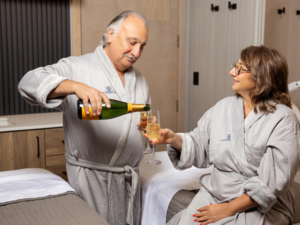 A mature couple in plush robes cheerfully pours champagne while relaxing in a luxurious spa setting.