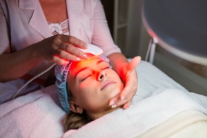 A patient undergoing LED light therapy facial treatment, with a protective hair cap and eyeshades, while a clinician holds a glowing red device over the face.