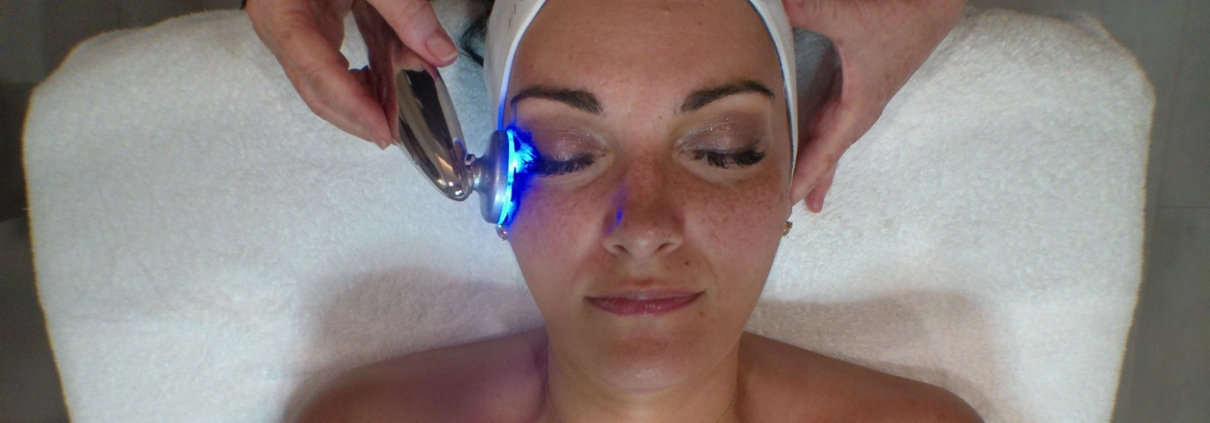 Woman lying on a treatment bed with an LED device applied to her cheek, wearing a Five Elements Spa headband.
