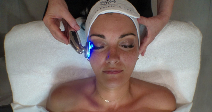 Woman lying on a treatment bed with an LED device applied to her cheek, wearing a Five Elements Spa headband.