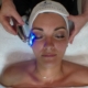 Woman lying on a treatment bed with an LED device applied to her cheek, wearing a Five Elements Spa headband.